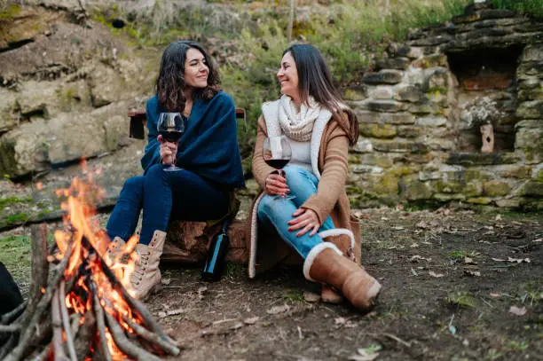young happy women talking together while drinking glass of red wine. Females warming next to the fire. Campfire, outdoors activities concept.