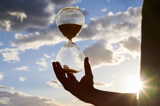 istock man's hand holding backlited hourglass with a half cloudy sky in the background 1298360668