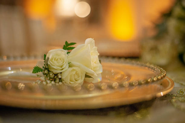 istock Three white roses on gold charger on table at wedding reception with warm bokeh background 1298361435