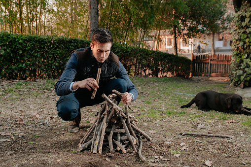 Portrait young man preparing pile of wood to turn on fire. Dog on background. Camping, natural lifestyle concept.