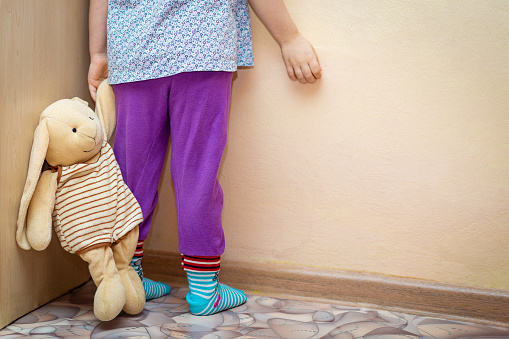 A small child stands in the corner, grounded, holding a stuffed bunny by the ear. Selective Focus