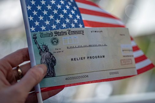 Washington, DC, USA - July, 8, 2020:  COVID-19 economic Stimulus check (American rescue program) on blurred USA flag background. Relief program concept.