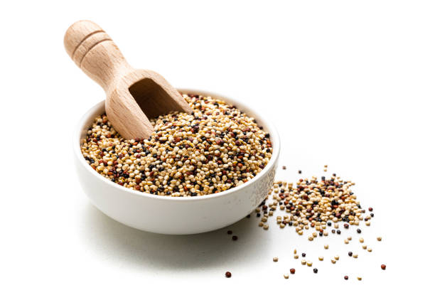 Close up view of a white bowl filled with a mixture of red, black and white quinoa seeds isolated isolated on white background. A wooden serving scoop is inside the bowl and some seeds are scattered on the table. High resolution 42Mp studio digital capture taken with Sony A7rII and Sony FE 90mm f2.8 macro G OSS lens
