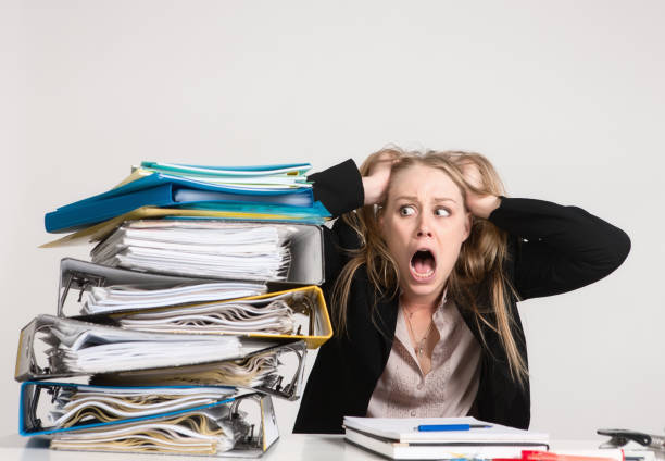 Beautiful young blonde woman looks daunted by stack of files Young woman in her 20s faces the prospect of a mountain of work. overworked funny stock pictures, royalty-free photos & images