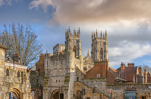 The west facing towers of York Minster rise over the roofs of some houses.  An ancient city gateway is in the foreground and a cloud filled sky is above.