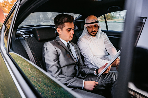 Emirati executive man and a western businessman sitting on back seat of a car and talking about work. Doing some paperwork and planning.