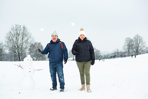 Couple poses standing next to a snowman in a park during a cold winter day. They are throwing snowballs.