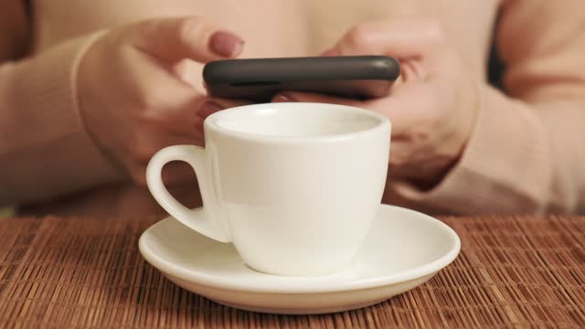 A woman with a mobile phone in her hands and drinking coffee in a white cup.