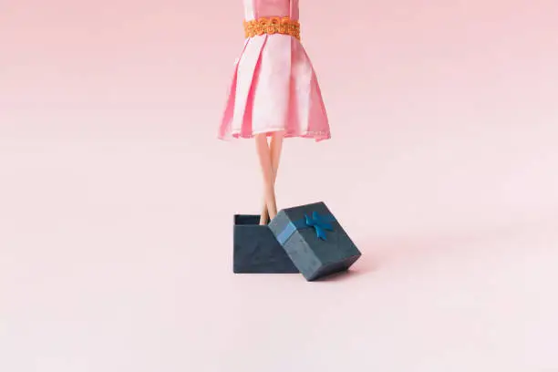 Photo of A woman in a pink dress comes out of a blue gift box. Pink background.