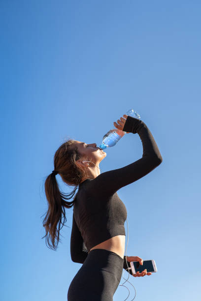 istock Thirsty woman drinking water from plastic bottle, resting after jogging, holding mobile smartphone, listens to music. Hot day. 1298428999