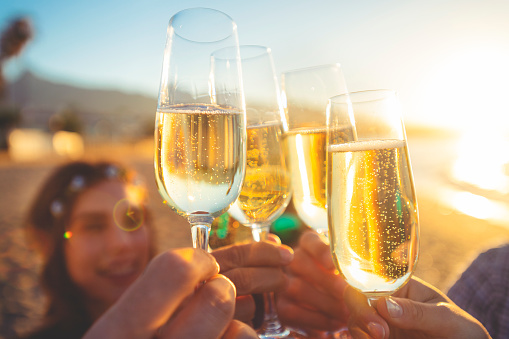 Group of friends making a champagne toast on the beach. The party is on the beach at sunset. Everyone is looking cool, laughing and smiling.