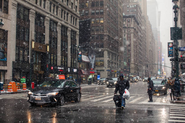 Adult man with travel suitcase crosses street. Goes not along the pedestrian crossing, but along the roadway. Snow continues to fall and snowflakes are visible flying in the air. The sidewalk and road are covered with snow. The carriageway is wet and glossy. Photo from a middle of the intersection headed to North of 7th Avenue. Penn station area. Some yellow NYC taxis and cars to hire on the photo, including the foreground. Traffic lights, trucks and cars visible on the photo. High-rise buildings on the left and right. In the distance, skyscrapers are in snowy haze. Snow continues to fall and snowflakes are visible flying in the air. The lighting of Time Square can be seen far behind. Slippery. Very bad weather. Evening. March 21, 2018. NYC. Midtown Manhattan. New York. USA
