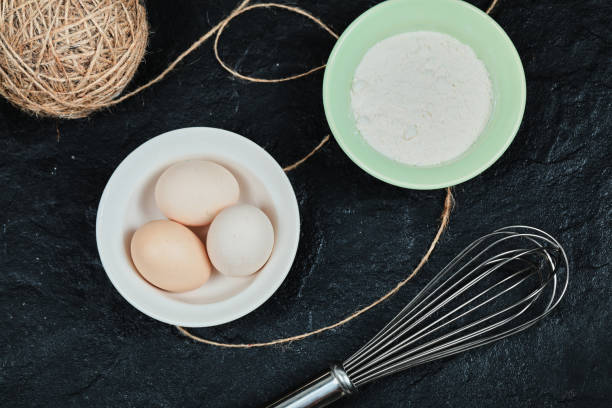 istock Chicken eggs on ceramic plate and a bowl of flour on dark table with whisker 1298433994