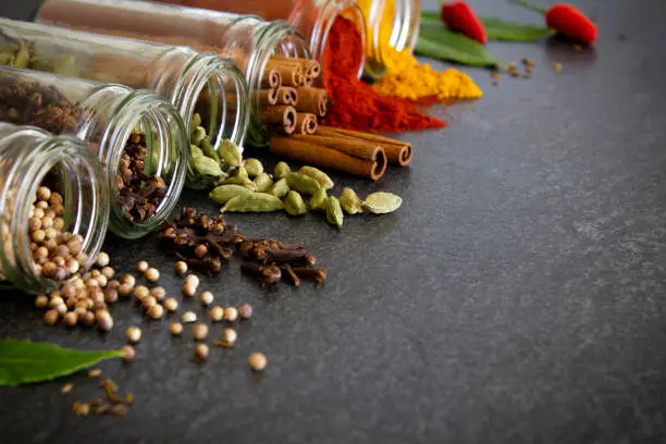 A selection of dried spices and herbs spilling out from glass jars onto a dark slate background. 
Coriander seed, cloves, cardamom, cinnamon sticks, chilli powder, turmeric and fresh bay leaves. 
Low angle view with copy space to the right of the image.  
Close up.