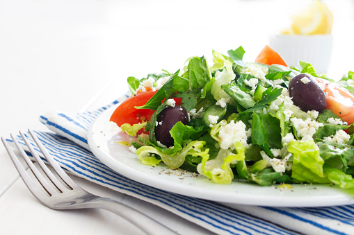 A fresh Greek salad with feta cheese, black olives, herbs  and salad dressing served on a white table with a blue striped napkin. 
Low angle view with  copy space to the top left of the image.