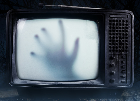 Horror photo of an old black  scary haunted tv set with ghost hand on a screen, standing on dark foggy background.