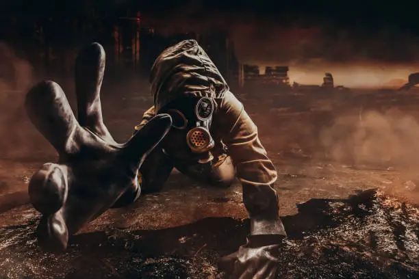 Photo of a dying stalker in jacket and gloves in damaged gas mask with filter reaching out his hand to camera on destructed apocalyptic wasteland city background.