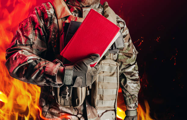 istock Soldier in uniform holding closed red book on burning flame background. 1298444943