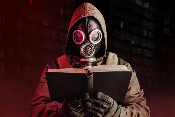 Photo of stalker soldier in soviet gas mask holding opened old book on dark background.