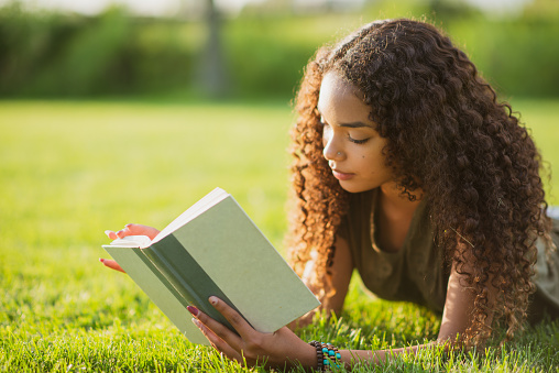 A beautiful teenager of African ethnicity is lying on the grass outdoors. She is reading a book.