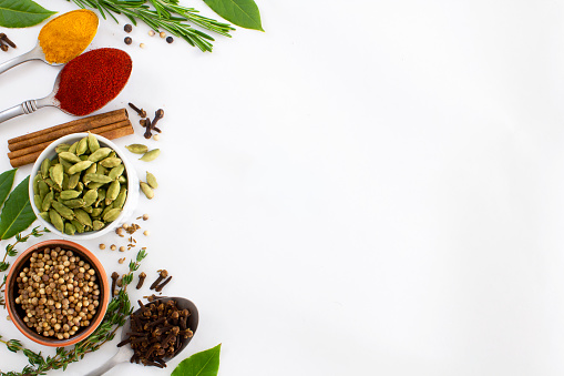 A selection of dried spices and herbs on a white  background. \nCoriander seeds, cloves, cardamom, cinnamon sticks, chilli powder, turmeric and fresh bay leaves. \nShot from above with copy space on the right of the image. Flat lay.