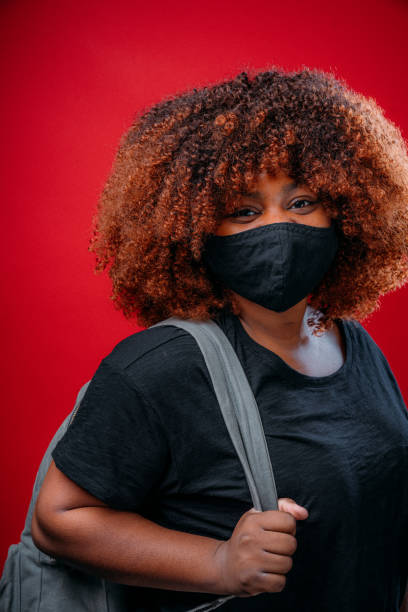 istock Young African American Woman Smiling Under a Cloth Protective Face Mask Wearing a Backpack Getting Ready for School in COVID-19 1298494182