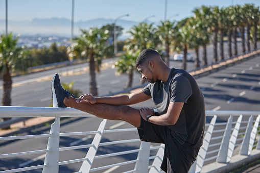 Shot of a sporty young man stretching his legs while exercising on a bridge in the city