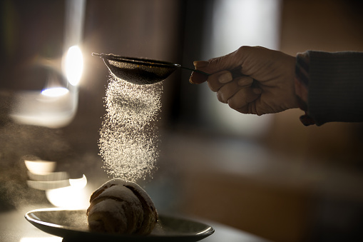 Close up of unrecognizable woman sprinkling powdered sugar from a sieve on a croissant.