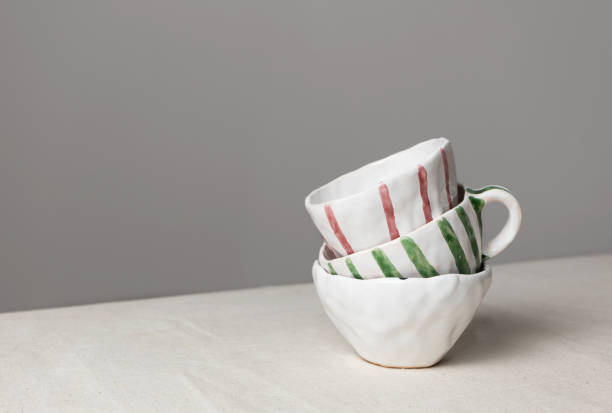 istock White ceramic cups with colorful stripes stack on table with linen tablecloth and gray wall background. Close-up handmade ceramic, horizontal 1298524660