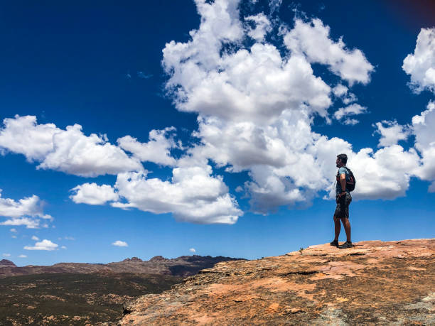 Side view Hiker Trail runner mid age Caucasian male with active rucksack on back Hands on hips standing on rocky mountain summit looking out beautiful cloud and mountain backdrop in the Cederberg Mountain Wilderness in summer South Africa