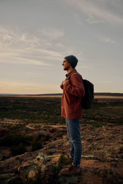 Successful hiker with bag and cap standing on mountain cliff looking at morning sunrise
