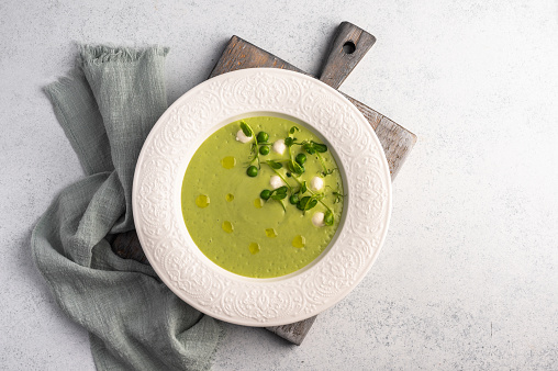 Homemade soup puree of green peas, coconut milk, mini mozzarella cheese in white plate wth napkin on wooden board, top view