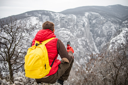 A man on vacation hiking. Wearing warm clothes and a backpack. He took a break to rest and enjoy the beautiful scenery. Winter day. The mountains are covered with snow.