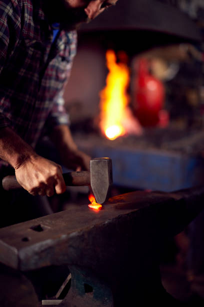 istock Male Blacksmith Hammering Metalwork On Anvil With Blazing Forge In Background 1298555395
