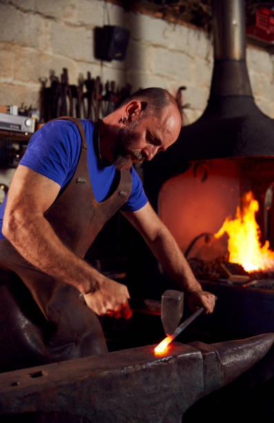 istock Male Blacksmith Hammering Metalwork On Anvil With Blazing Forge In Background 1298556510