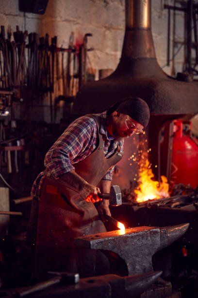 istock Male Blacksmith Hammering Metalwork On Anvil With Blazing Forge In Background 1298556520