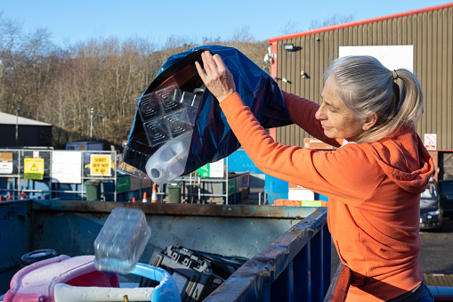 Woman dropping off her home recycling at a recycling hub in the North East of England.  She is tippng a bag of mixed recycling into a container.