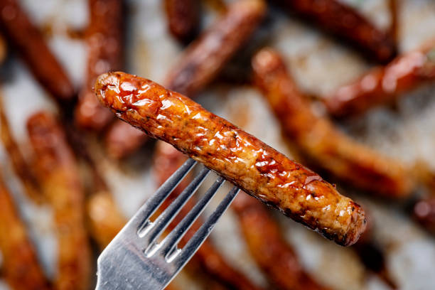 Close up of a grilled sausage on a fork. With a grill pan of some sausages cooking in the background. Colour, horizontal format. Could be used to illustrate diet or even global warming as meat production is a big contributor to greenhouse gases.