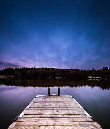 Long exposure image depicting a calm and peaceful lake with a perfect reflection, and a wooden jetty protruding into the distance with a forest on the other side of the lake.