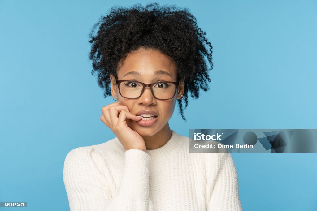 Shy awkward Afro woman wear glasses biting finger feeling embarrassed, confused and nervous. Studio. Close up studio portrait of shy awkward young Afro American woman wear glasses white sweater, biting nails feeling embarrassed, confused and nervous, looking at camera, isolated on blue background. Embarrassment Stock Photo