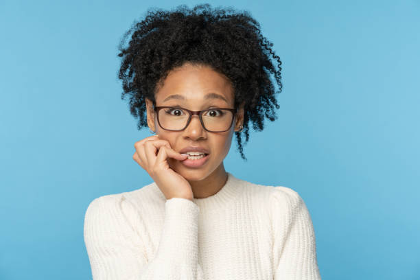 Shy awkward Afro woman wear glasses biting finger feeling embarrassed, confused and nervous. Studio. Close up studio portrait of shy awkward young Afro American woman wear glasses white sweater, biting nails feeling embarrassed, confused and nervous, looking at camera, isolated on blue background. embarrassment stock pictures, royalty-free photos & images