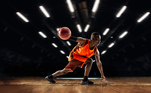 Team supporting. African-american young basketball player in action and motion in flashlights over dark gym background. Concept of sport, movement, energy and dynamic, healthy lifestyle. Arena's drawned.