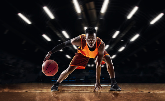 Ready to jump. African-american young basketball player in action and motion in flashlights over dark gym background. Concept of sport, movement, energy and dynamic, healthy lifestyle. Arena's drawned.