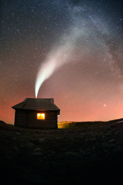 The starry sky at a mountain shelter in the Alps is photographed at long exposure. Thick white smoke comes from the stove in which the wood is burning