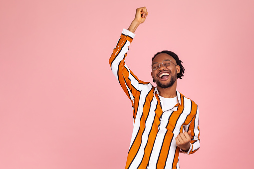 Excited positive bearded afro-american man in stylish eyeglasses and striped shirt dancing, raising hands up and having fun, good mood. Indoor studio shot isolated on pink background.