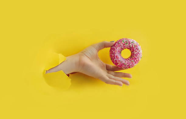 istock Unrecognizable hand holds delicious round glazed donut with sprinkle in torn yellow bright paper hole, popular food blog content, selective focus 1298589310