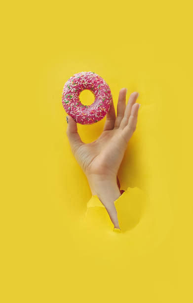 istock Unrecognizable hand holds delicious round glazed donut with sprinkle in torn yellow bright paper hole, vertical popular food blog content, selective focus 1298589324