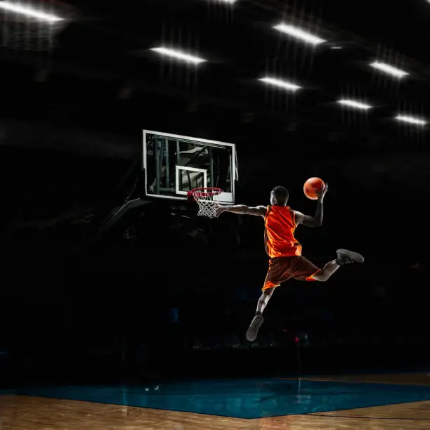 Photo of African-american young basketball player in action and flashlights over gym background. Concept of sport, movement, energy and dynamic, healthy lifestyle.