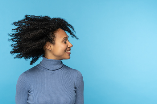 Happy African woman smiling, has good mood, dancing with her black curly hair flying, isolated on studio blue background. Excited Afro American girl with flying hairstyle. Positive life concept.