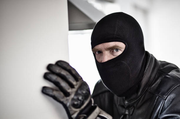 Scary man masked by black balaclava and clad in leather looks furtive and startled: possible burglar Ominous balaclava-clad man dressed all in black, inside a building. clad stock pictures, royalty-free photos & images